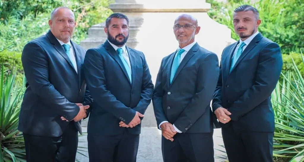 Quatre porteurs funéraires en costume sombre et cravate turquoise posant devant un monument